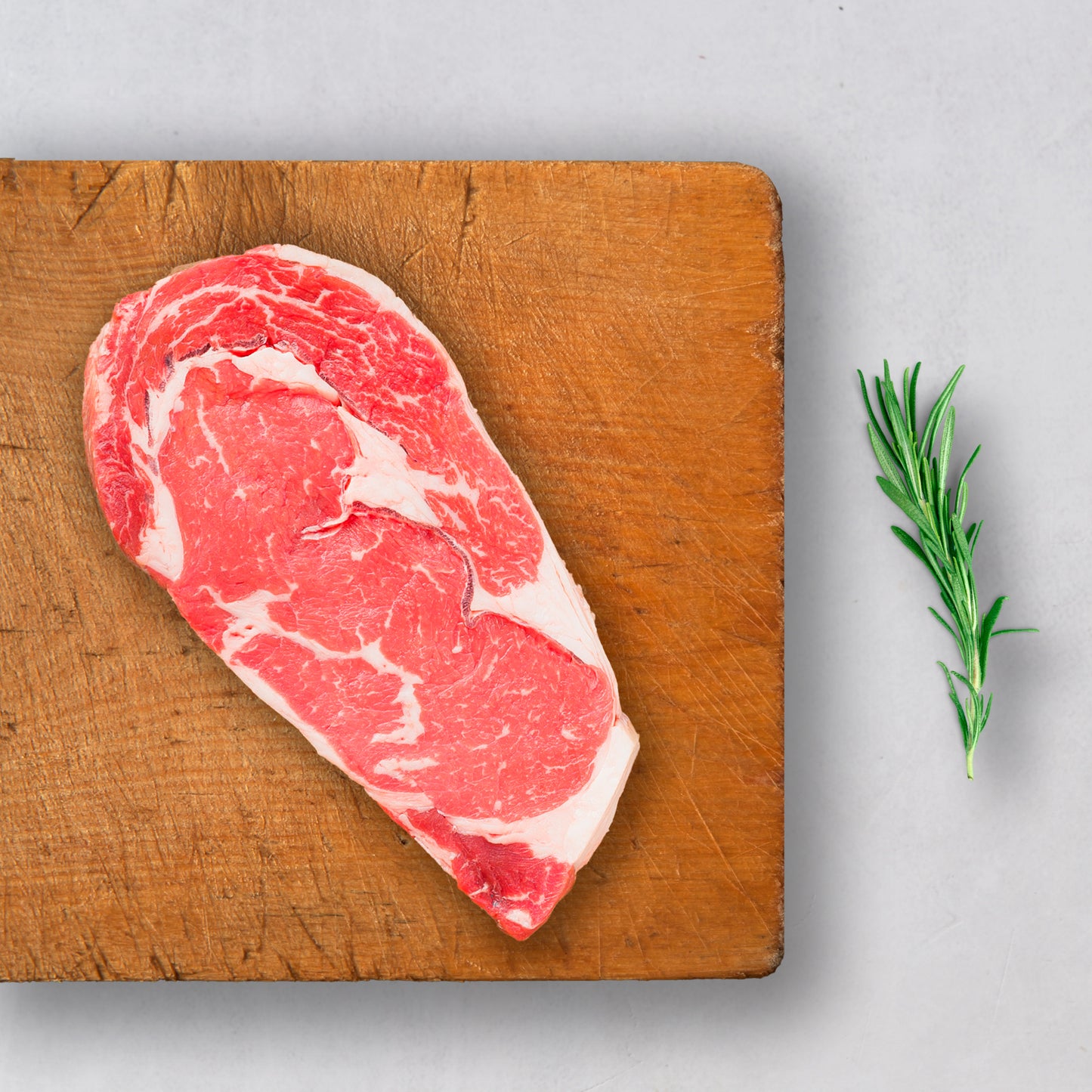 A Ninth & Prime Boneless Ribeye steak with rich marbling sits on a wooden cutting board beside fresh rosemary, displayed on a light gray surface.