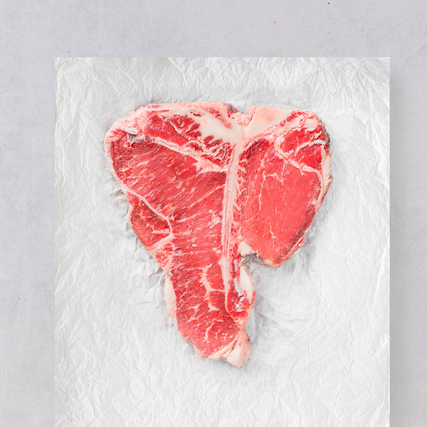 A raw Ninth & Prime Porterhouse steak, showcasing tenderloin and marbled fat, sits on crumpled aluminum foil with a light gray background.