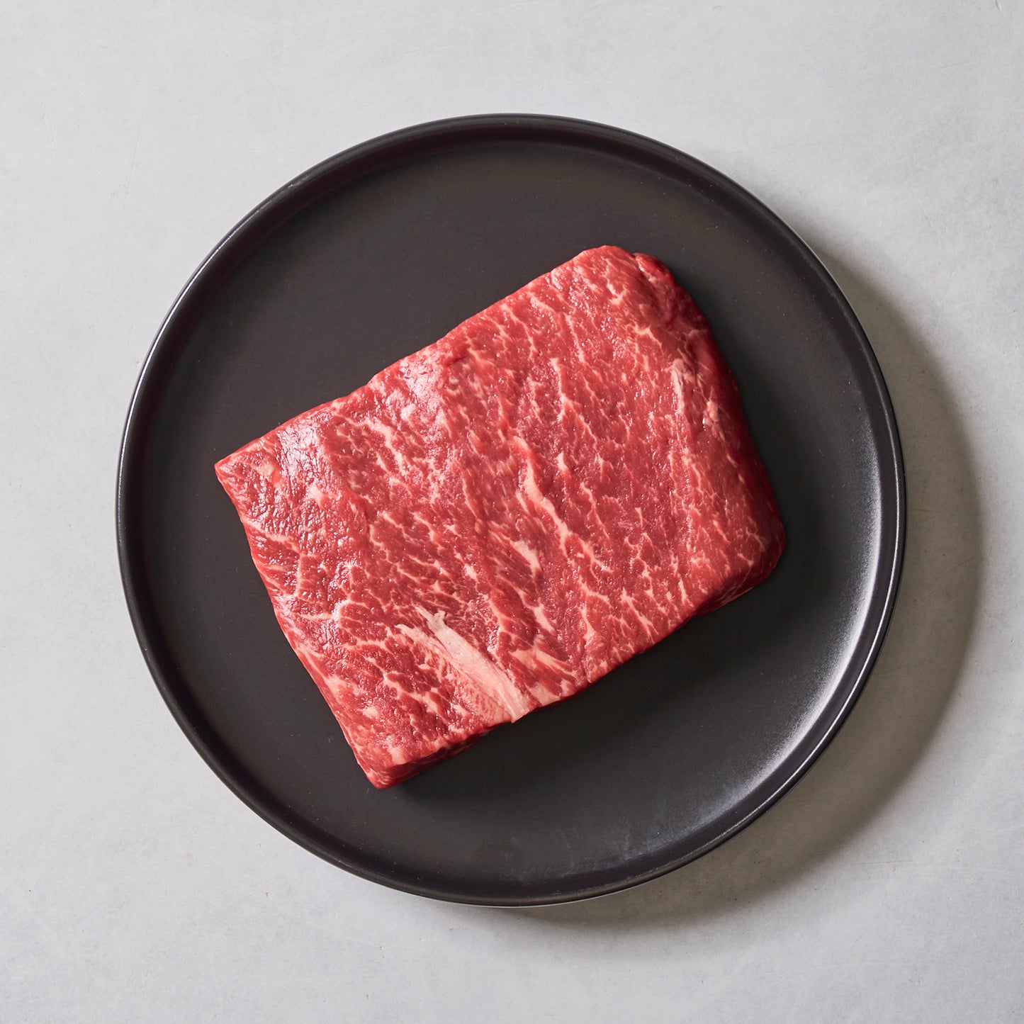 The Ninth & Prime Flat Iron Steak sits on a round black plate against a light gray background, showcasing its marbling and promising rich, beefy flavor in every bite.