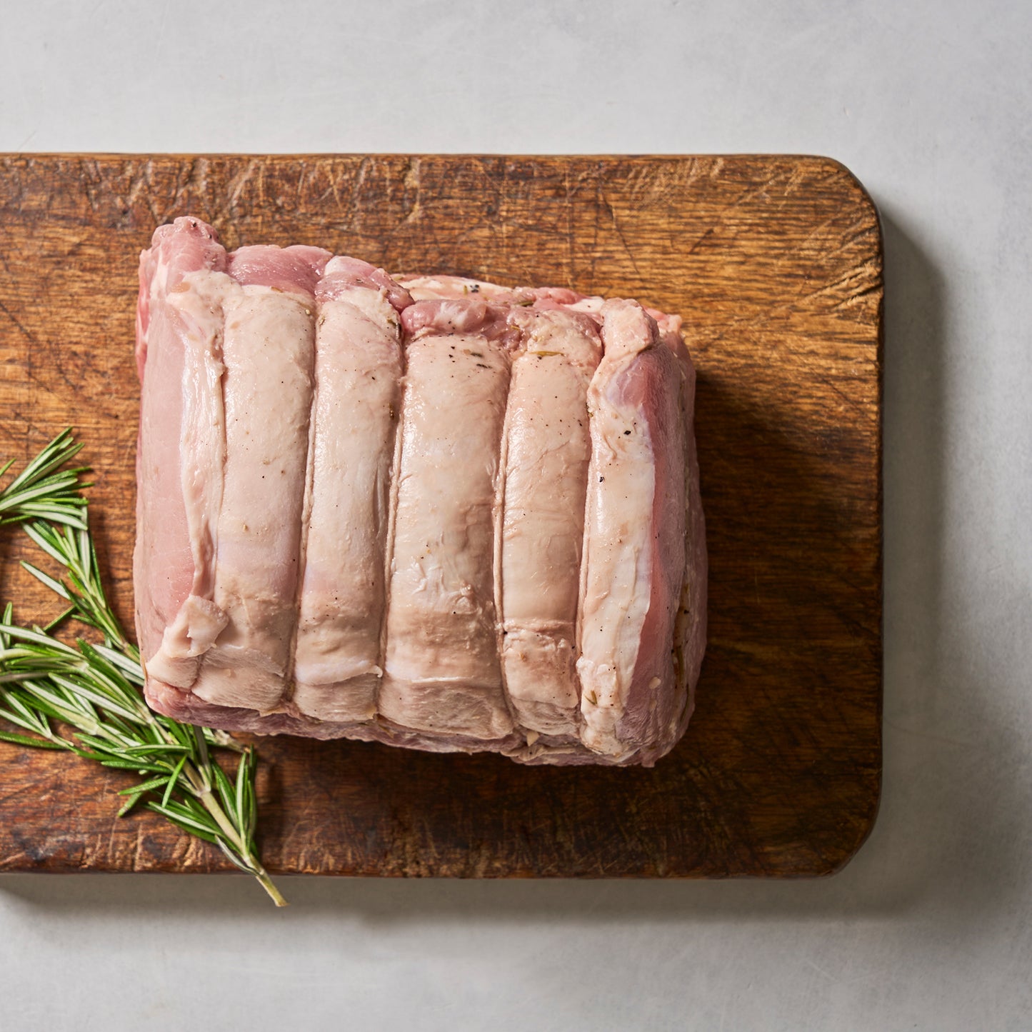 Ninth & Prime Porchetta Roast, a raw boneless pork roast tied with string and accented by herbs and fresh rosemary, sits on a wooden cutting board ready for roasting.