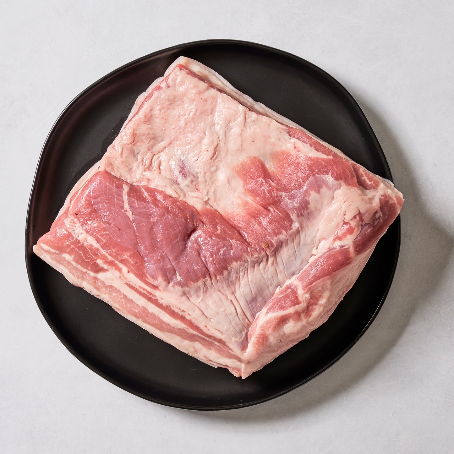 A large, raw Ninth & Prime Pork Belly with distinct fat and meat layers rests on a round black plate over a light surface, ready to be slow-cooked into a gourmet dish.