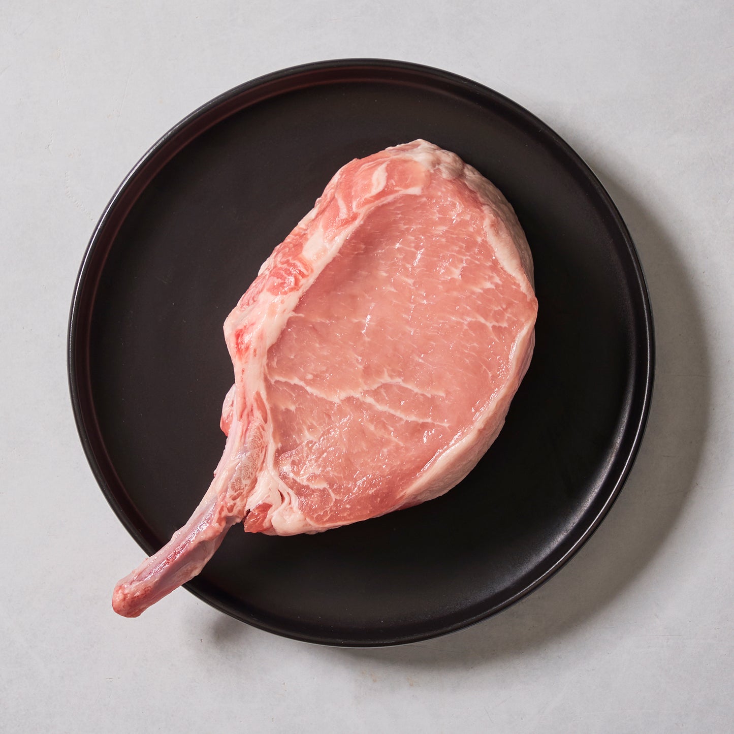A raw Ninth & Prime Pork Rib Chop, prized for its flavor and tenderness, rests on a round black plate set against a light gray background.