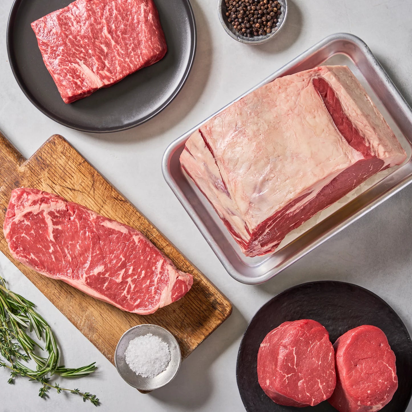 Our Prime Picks by Ninth & Prime features assorted raw beef cuts like Filet Mignon and NY Strip, artfully arranged with salt, peppercorns, and fresh herbs on plates and a wooden board atop a light surface.