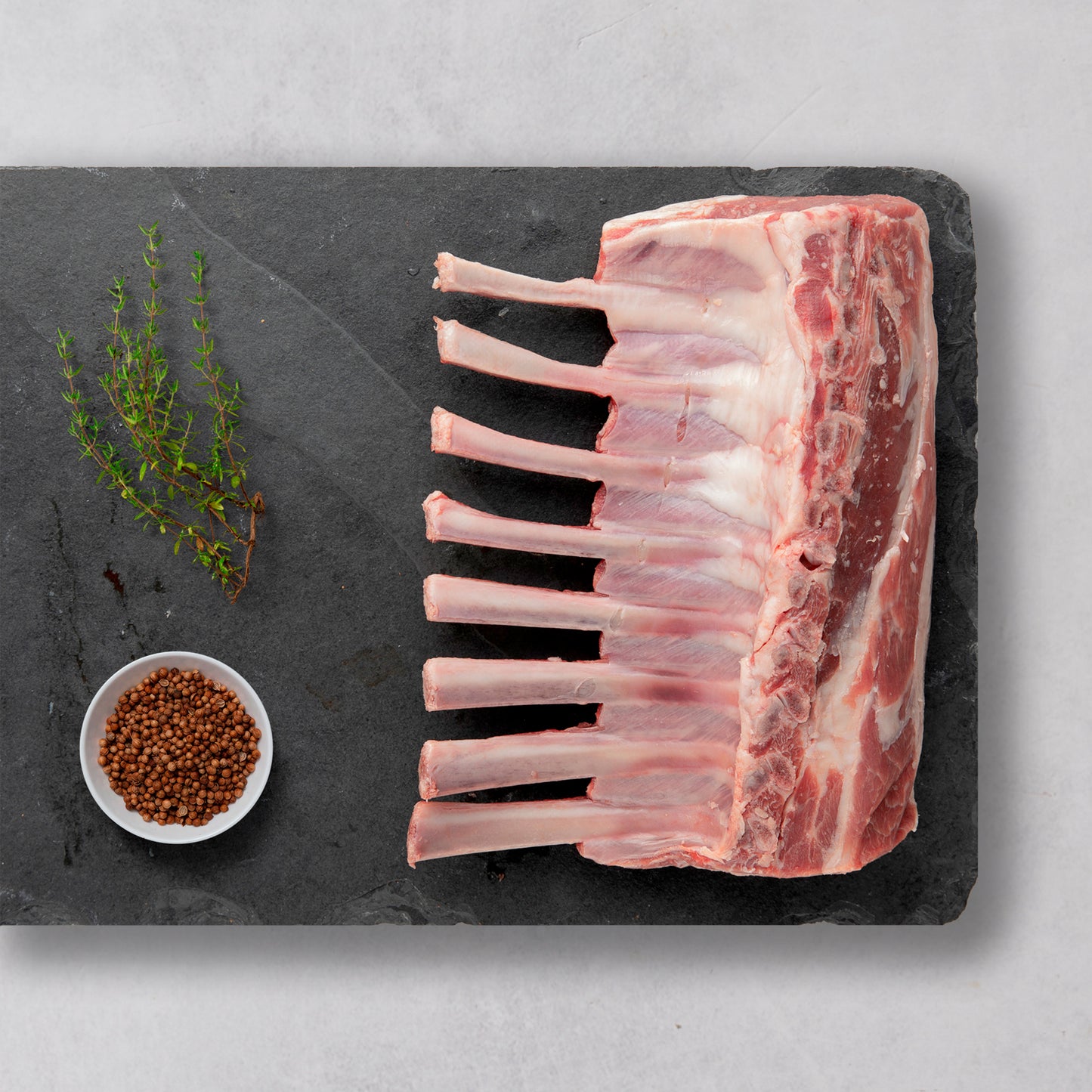 Ninth & Prime Grass-Fed Rack of Lamb, featuring eight ribs, is artfully displayed on a dark slate board with fresh thyme and whole coriander seeds—highlighting this premium cut on a light gray surface.