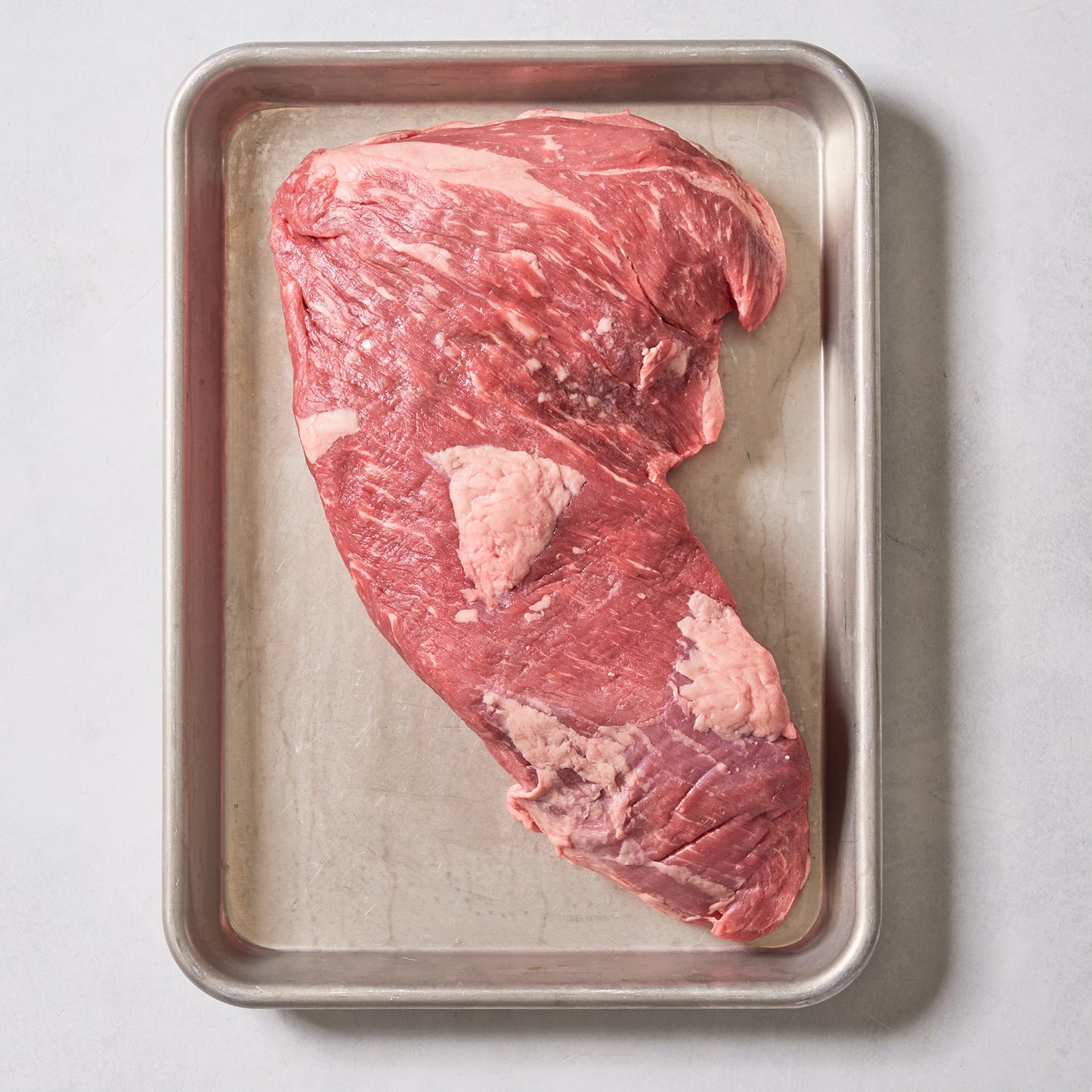 A raw Ninth & Prime Tri-Tip Steak sits on a rectangular metal tray against a light background, displaying visible marbling and patches of fat that highlight its juicy tenderness and bold flavor.