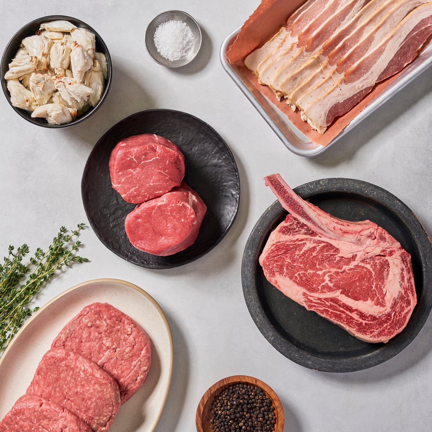 A top-down view of the Yacht Box by Ninth & Prime shows colossal crab meat, bacon, filet mignon steaks, bone-in ribeye, and ground beef patties on plates with bowls of salt, pepper, and fresh thyme.