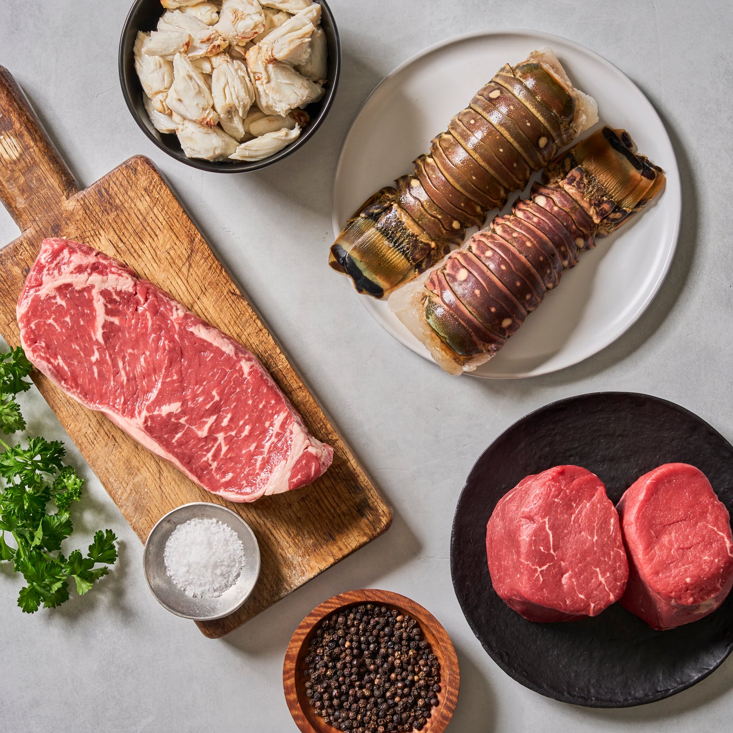 A top-down view of Ninth & Prime's Surf & Turf, featuring raw steak, Cold Water Lobster Tails, filet mignon, Colossal Crab Meat, parsley, salt, and whole black peppercorns arranged on a table.