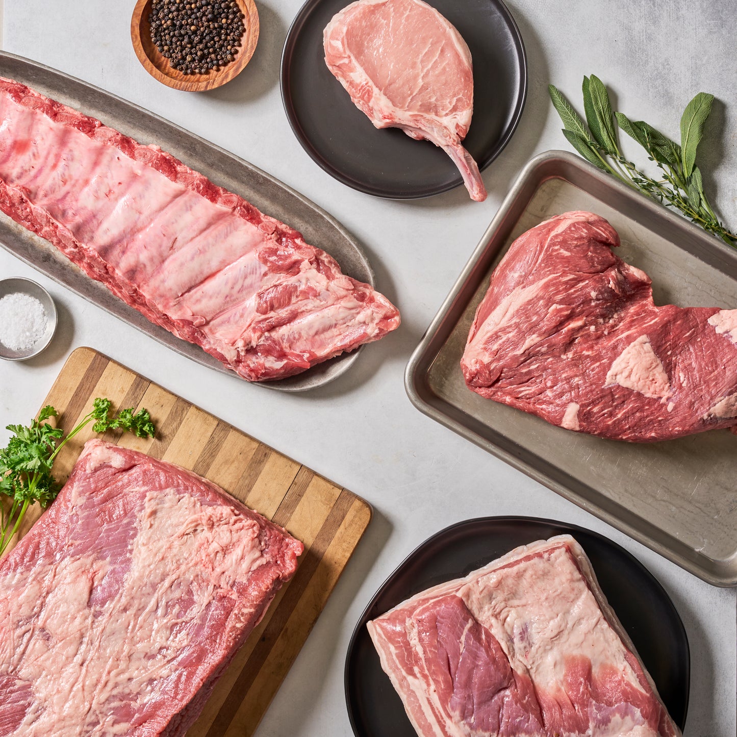 Ninth & Prime Que It Up features assorted raw pork cuts on plates, trays, and a cutting board with herbs, peppercorns, and salt, all set on a light-colored surface.