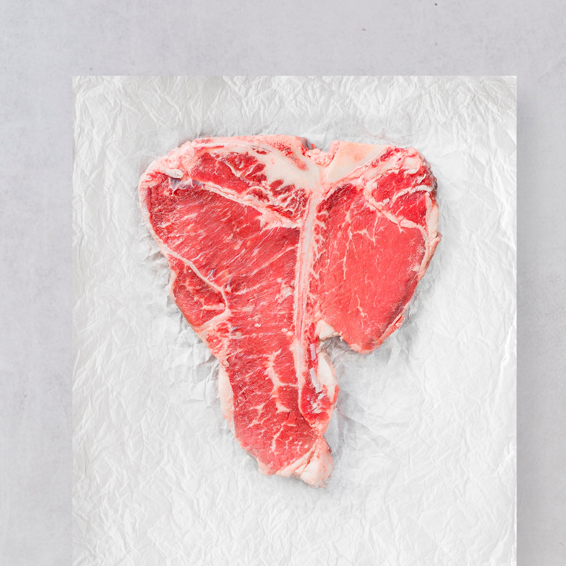 A raw Ninth & Prime Porterhouse steak, showcasing tenderloin and marbled fat, sits on crumpled aluminum foil with a light gray background.