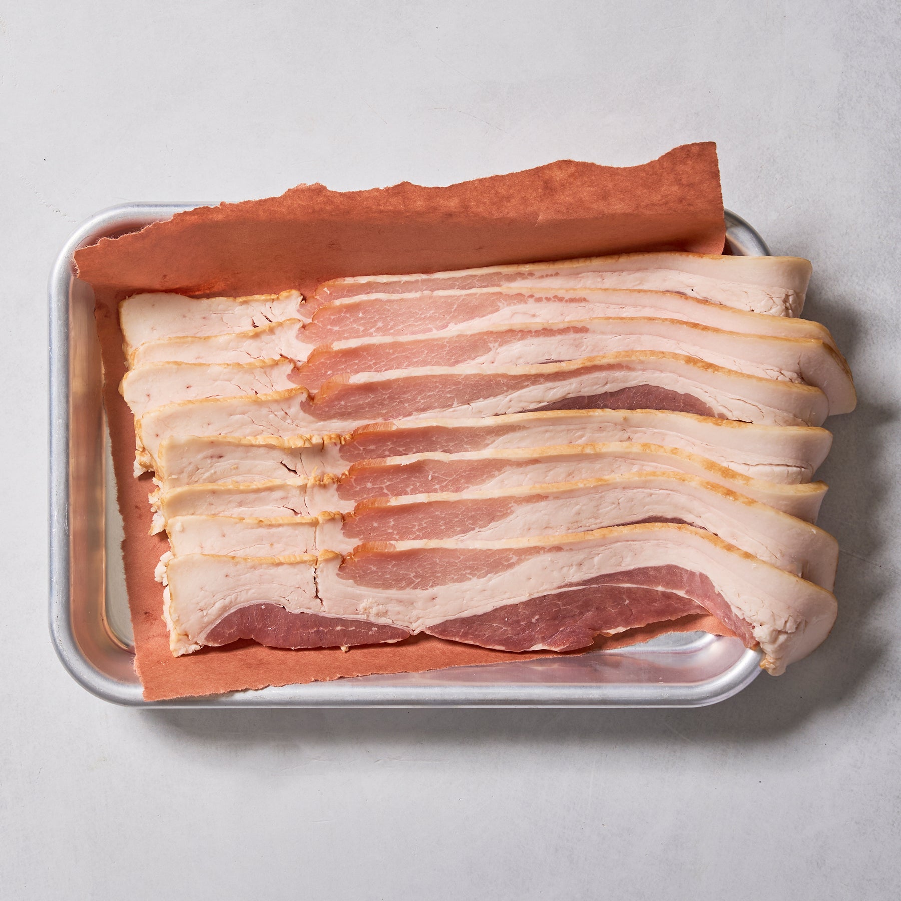 Several strips of Ninth & Prime Millionaire Thick Cut Bacon are neatly arranged on a metal tray lined with brown parchment paper, set against a light-colored background.
