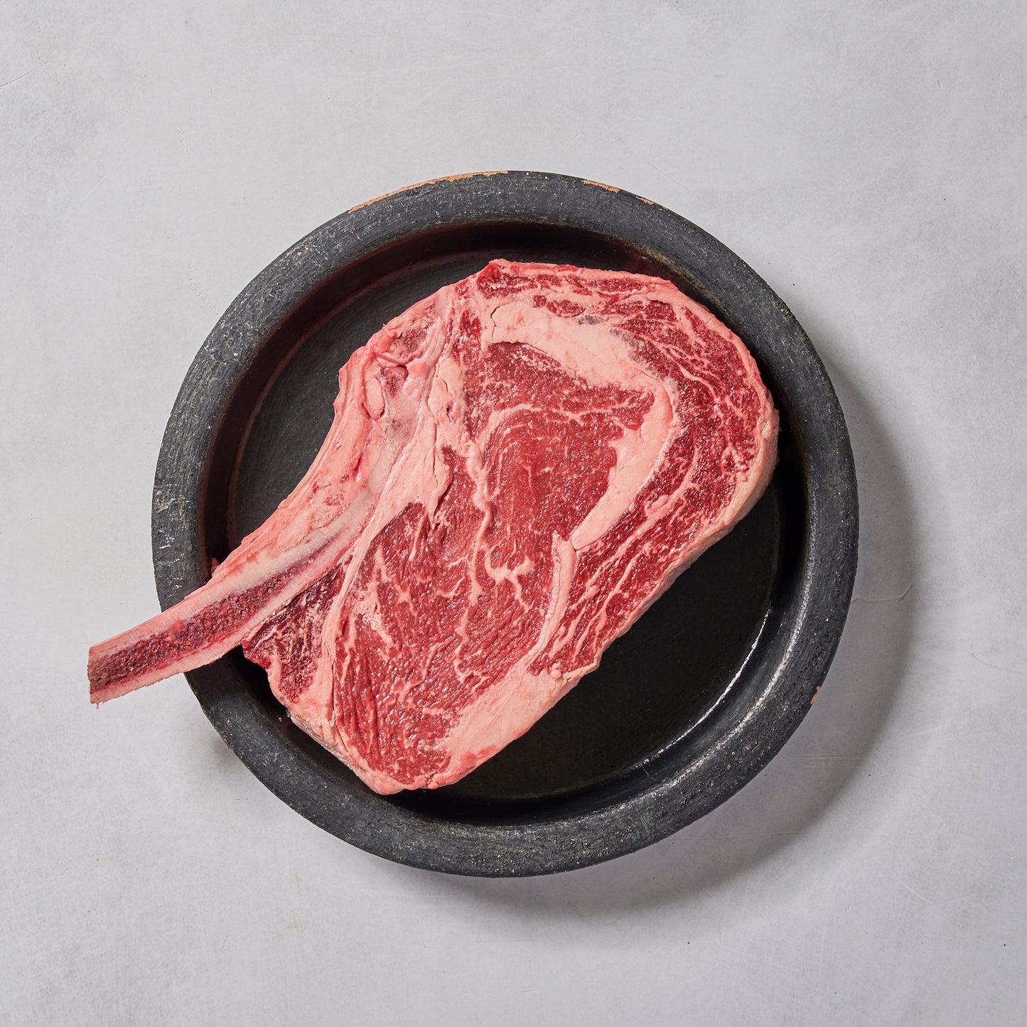 Raw bone-in ribeye steak with marbled fat placed on a round black plate against a light gray background