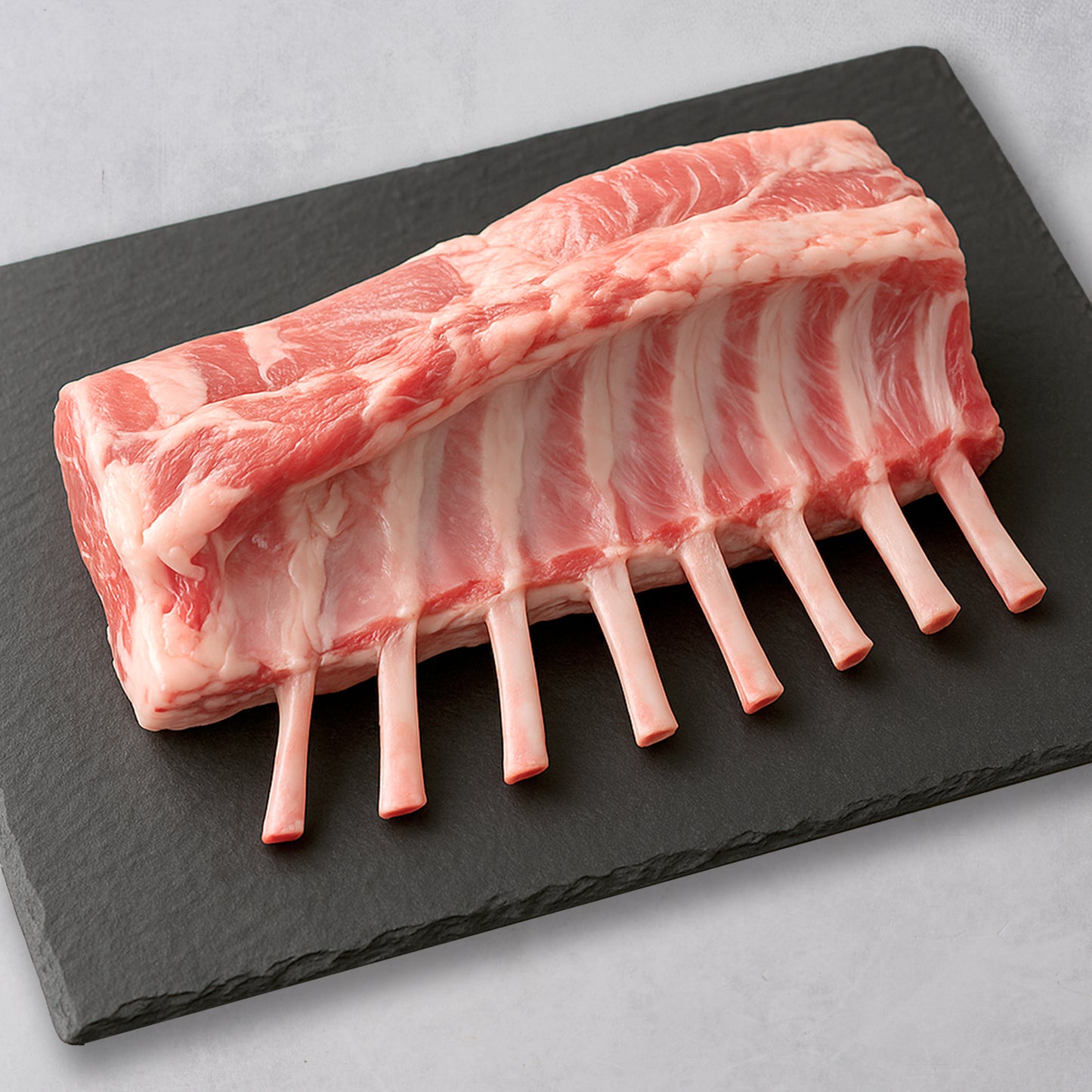 Ninth & Prime Domestic Rack of Lamb with eight exposed rib bones is a premium cut, elegantly displayed on a black slate board against a light gray backdrop—a gourmet choice for refined palates.