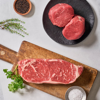 A marbled USDA Prime NY Strip steak and filet mignon steaks from Ninth & Prime's "Be the Best Gifter" collection are displayed with parsley, coarse salt, thyme, and black peppercorns on a cutting board and black plate.