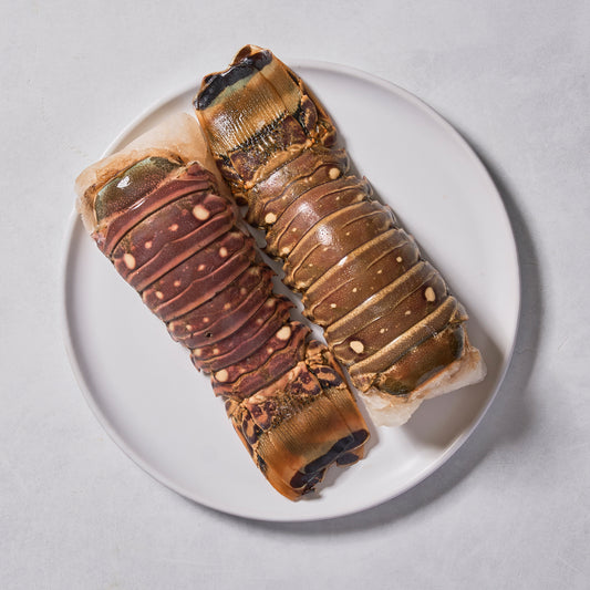 Two raw Ninth & Prime Lobster Tails with brown and tan shells rest side by side on a white plate, ready for an Atlantic-inspired seafood feast.