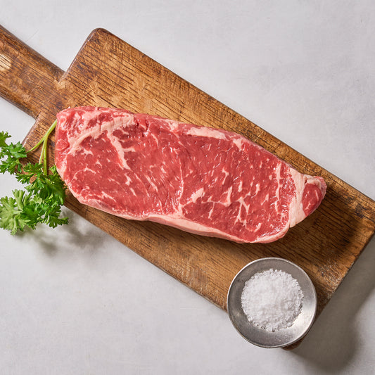 A raw Ninth & Prime NY Strip Steak sits on a wooden cutting board with parsley, accompanied by coarse salt in a small dish nearby on a light gray surface, evoking classic steakhouse vibes.