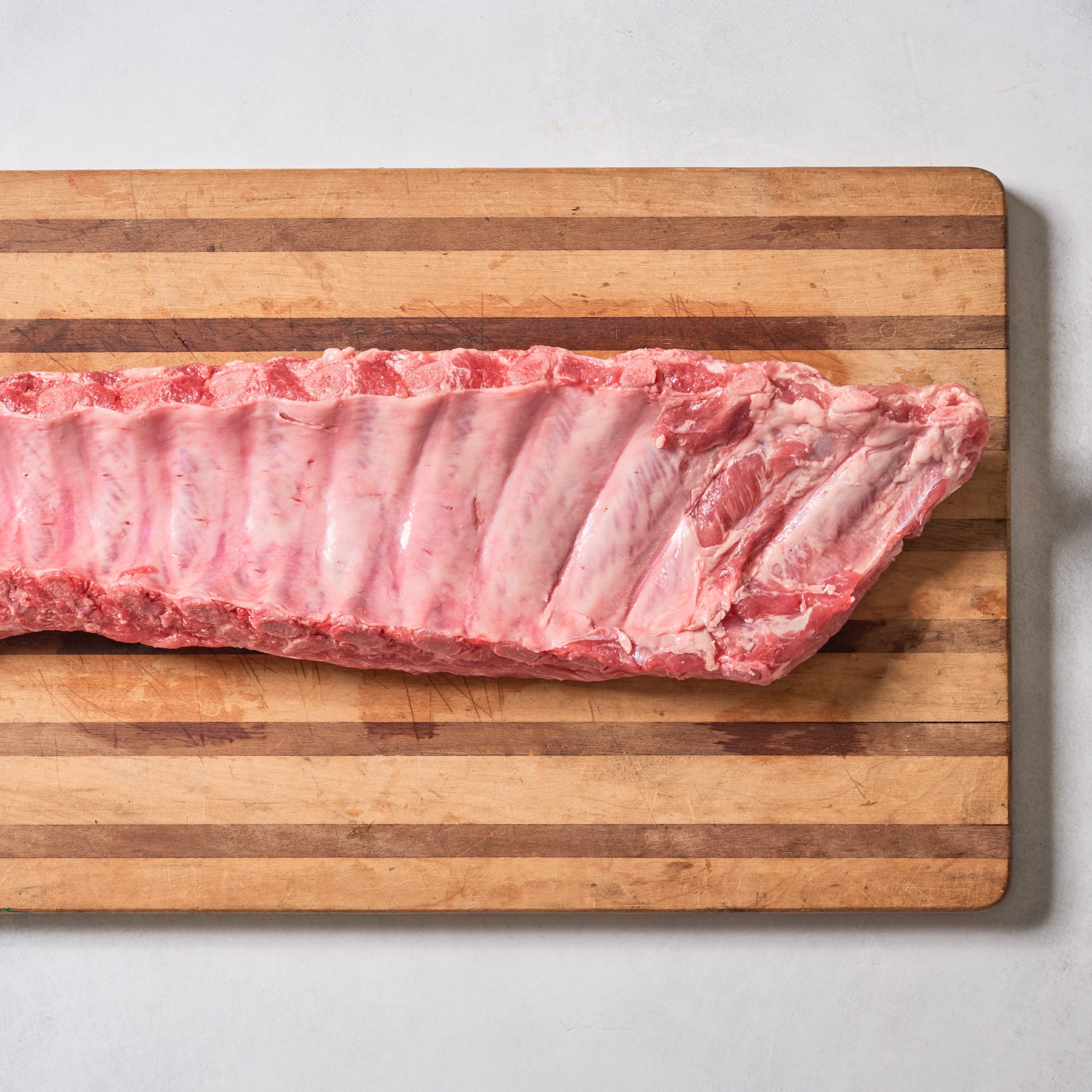 A rack of Ninth & Prime Baby Back Pork Ribs, an essential for barbecue, is placed horizontally on a striped wooden cutting board and photographed from above on a light surface.