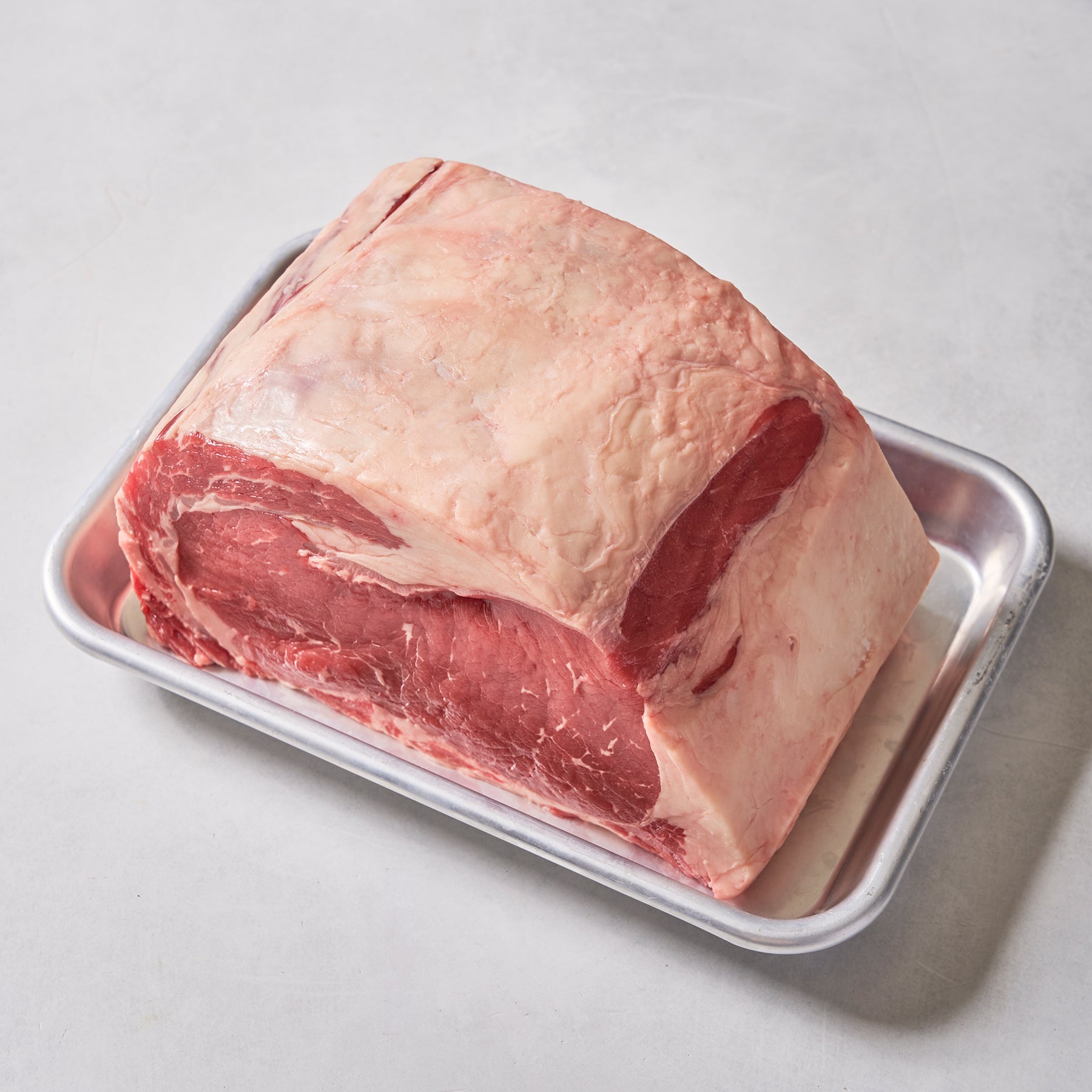 A Ninth & Prime Prime Rib Roast, richly marbled with a thick fat cap, rests on a rectangular metal tray against a light gray background, offering the promise of a luxurious dining experience.
