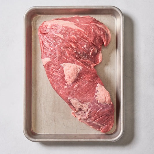 A raw Ninth & Prime Tri-Tip Steak sits on a rectangular metal tray against a light background, displaying visible marbling and patches of fat that highlight its juicy tenderness and bold flavor.