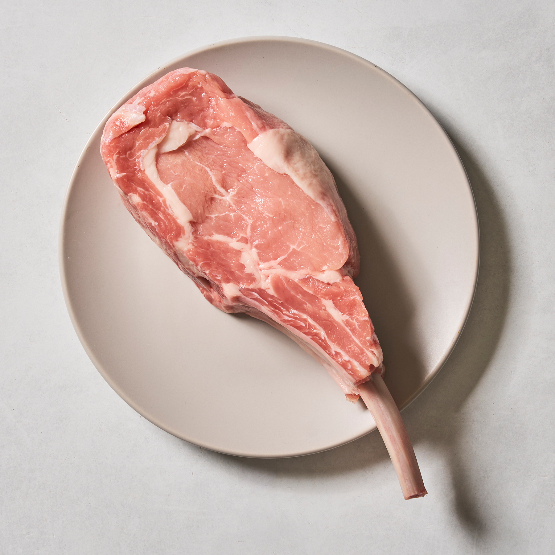 A Frenched Milk-Fed Veal Rib Chop by Ninth & Prime, featuring a long bone, is displayed on a plain beige plate against a light gray background, highlighting the premium quality and freshness of this exceptional veal cut.